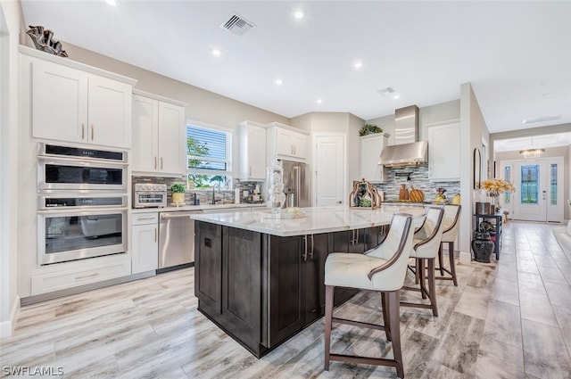 kitchen with appliances with stainless steel finishes, a center island, wall chimney range hood, backsplash, and light stone countertops