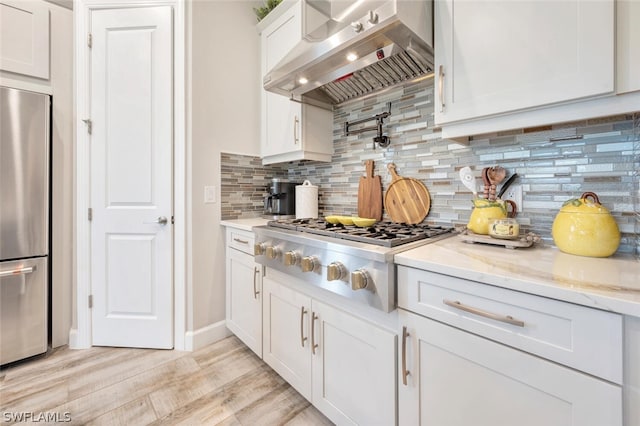 kitchen with wall chimney range hood, light wood-type flooring, backsplash, white cabinetry, and stainless steel appliances