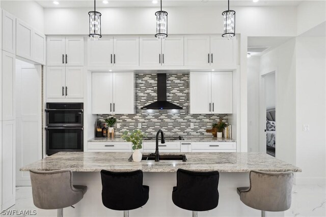 kitchen with white cabinetry, wall chimney exhaust hood, a center island with sink, and pendant lighting