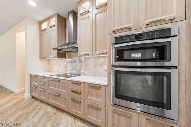 kitchen featuring double oven, wall chimney exhaust hood, tasteful backsplash, light wood-type flooring, and black electric cooktop