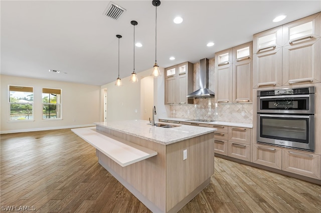 kitchen featuring a kitchen island with sink, double oven, wall chimney range hood, sink, and light hardwood / wood-style floors