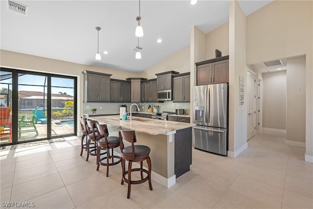 kitchen with decorative light fixtures, backsplash, appliances with stainless steel finishes, a center island with sink, and light stone counters