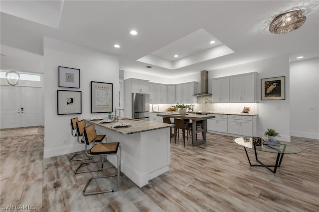 kitchen featuring light wood-type flooring, an island with sink, wall chimney exhaust hood, stainless steel refrigerator, and a breakfast bar area