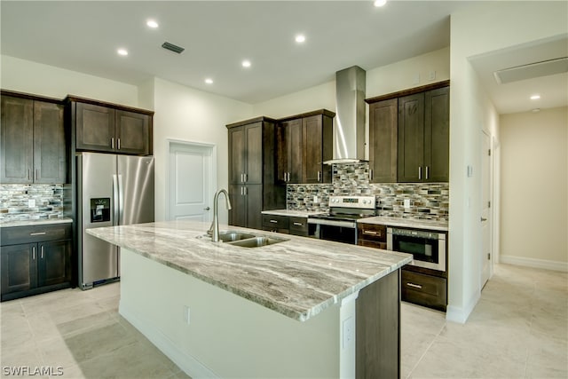 kitchen with backsplash, appliances with stainless steel finishes, a kitchen island with sink, wall chimney exhaust hood, and light stone counters