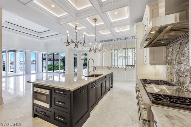 kitchen featuring wall chimney range hood, a notable chandelier, a center island with sink, sink, and pendant lighting