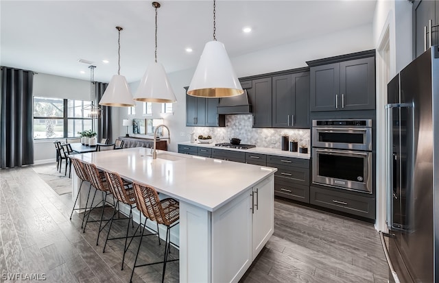 kitchen with a kitchen island with sink, hanging light fixtures, backsplash, stainless steel appliances, and sink