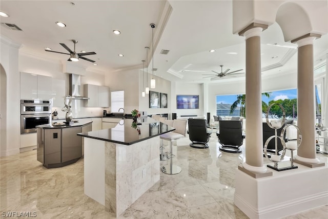 kitchen featuring hanging light fixtures, ceiling fan, wall chimney exhaust hood, and a center island with sink