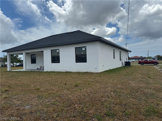 view of side of home with stucco siding, central AC unit, a lawn, and roof with shingles