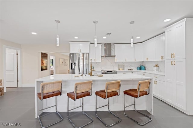 kitchen featuring white cabinetry, hanging light fixtures, an island with sink, stainless steel appliances, and wall chimney range hood