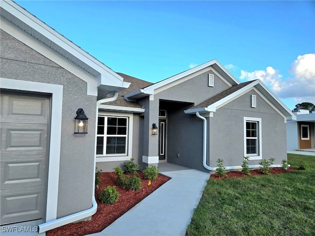 view of exterior entry with stucco siding, an attached garage, a lawn, and roof with shingles