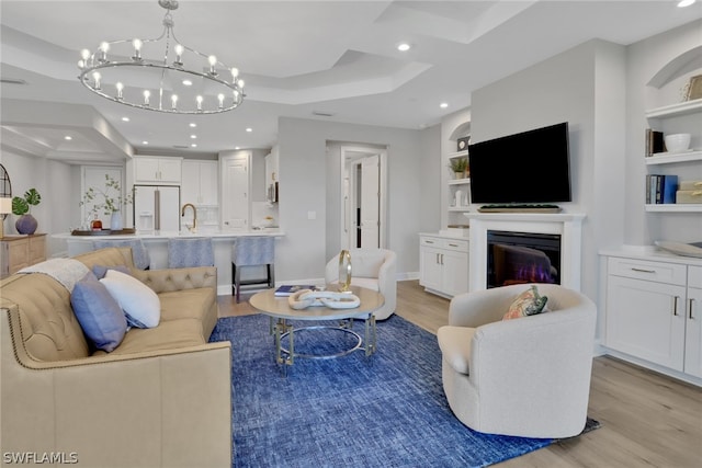 living room featuring a raised ceiling, light wood-type flooring, built in features, a notable chandelier, and sink