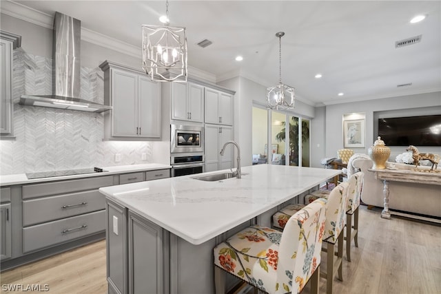 kitchen with gray cabinetry, appliances with stainless steel finishes, wall chimney range hood, a center island with sink, and sink