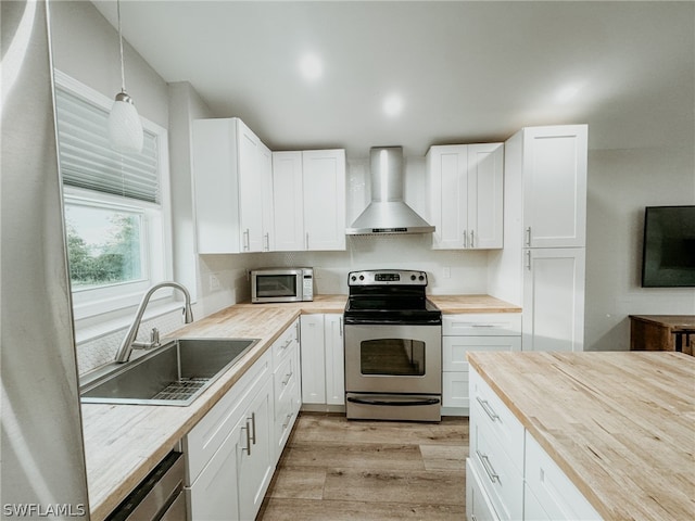 kitchen featuring wall chimney range hood, appliances with stainless steel finishes, butcher block countertops, decorative light fixtures, and sink