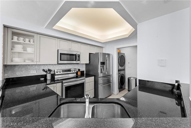 kitchen with stacked washer and dryer, sink, appliances with stainless steel finishes, a tray ceiling, and dark stone counters