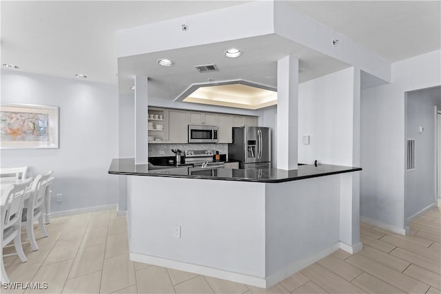 kitchen with gray cabinetry, a tray ceiling, kitchen peninsula, stainless steel appliances, and decorative backsplash