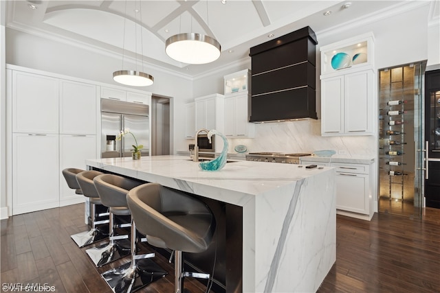 kitchen featuring white cabinetry, sink, built in refrigerator, hanging light fixtures, and a center island with sink