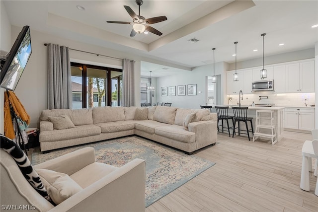 living room with ceiling fan, sink, a tray ceiling, and light hardwood / wood-style flooring