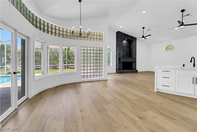 unfurnished living room with sink, a towering ceiling, light hardwood / wood-style floors, a large fireplace, and ceiling fan with notable chandelier