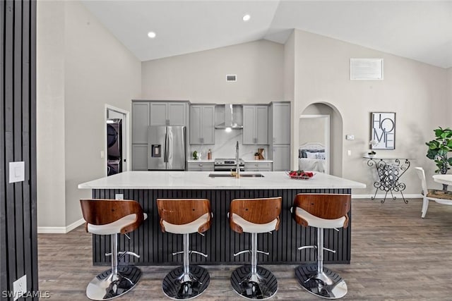 kitchen with gray cabinetry, wall chimney range hood, stainless steel refrigerator with ice dispenser, an island with sink, and a breakfast bar area