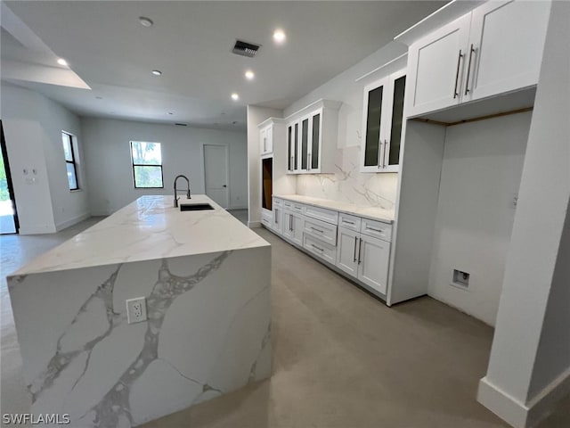 kitchen with light stone countertops, decorative backsplash, sink, white cabinetry, and an island with sink