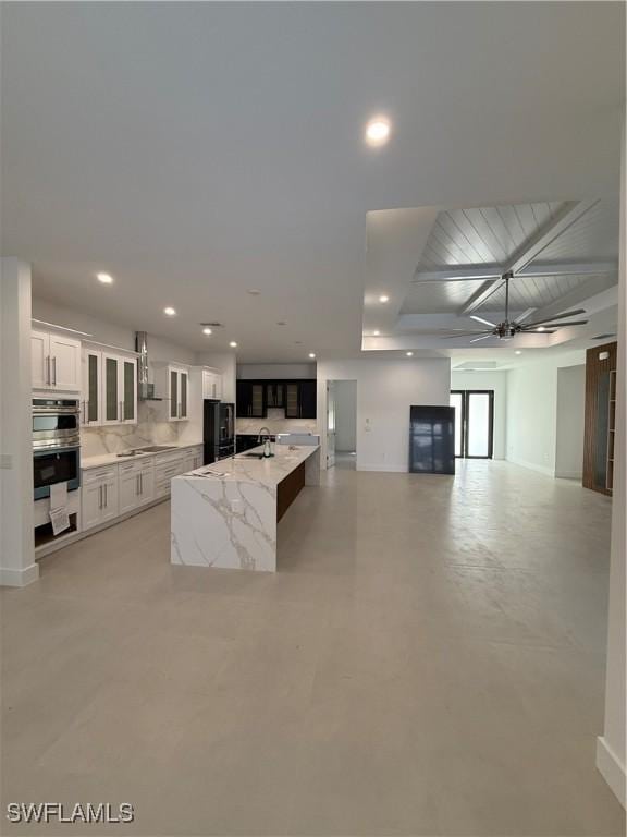 kitchen with white cabinetry, light stone counters, a center island with sink, black fridge, and wall chimney exhaust hood