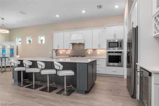 kitchen featuring premium range hood, an island with sink, white cabinets, and stainless steel appliances