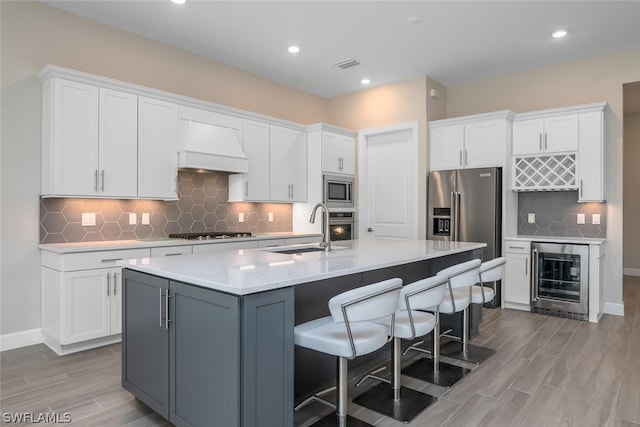 kitchen featuring custom exhaust hood, a kitchen island with sink, wine cooler, appliances with stainless steel finishes, and white cabinetry