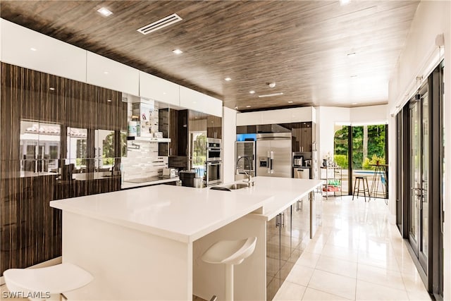 kitchen with an island with sink, stainless steel appliances, light tile flooring, wood ceiling, and wall chimney exhaust hood