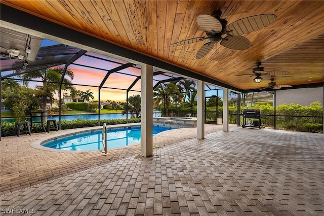 pool at dusk with an in ground hot tub, a water view, glass enclosure, grilling area, and a patio area