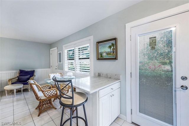 kitchen with light stone countertops, a wainscoted wall, light tile patterned floors, a kitchen breakfast bar, and white cabinets