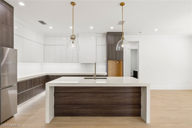 kitchen featuring stainless steel refrigerator, sink, an island with sink, decorative light fixtures, and white cabinets