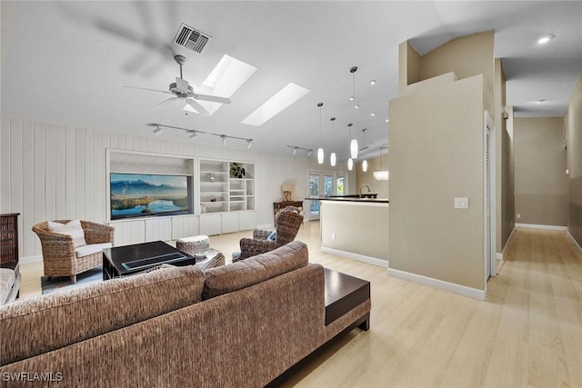 living room featuring built in shelves, rail lighting, ceiling fan, vaulted ceiling with skylight, and light hardwood / wood-style floors