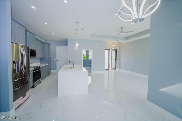 kitchen with ceiling fan with notable chandelier, sink, an island with sink, a tray ceiling, and stainless steel appliances