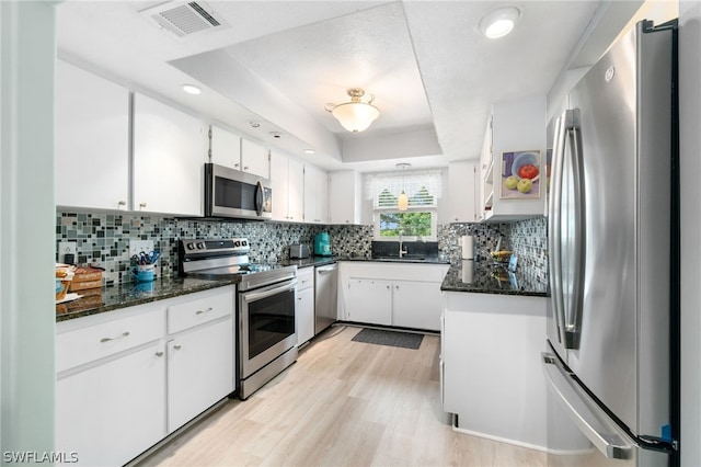 kitchen featuring light hardwood / wood-style flooring, stainless steel appliances, a raised ceiling, white cabinetry, and dark stone countertops