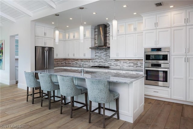 kitchen with wall chimney range hood, beamed ceiling, an island with sink, white cabinets, and appliances with stainless steel finishes