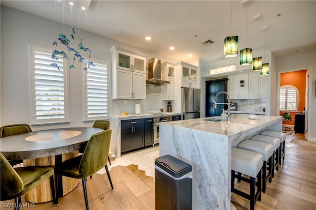 kitchen with light stone counters, hanging light fixtures, a center island with sink, wall chimney range hood, and white cabinets