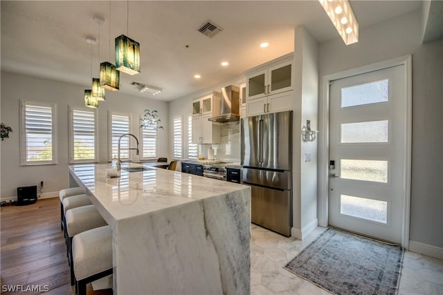 kitchen with appliances with stainless steel finishes, white cabinetry, an island with sink, light stone counters, and wall chimney range hood