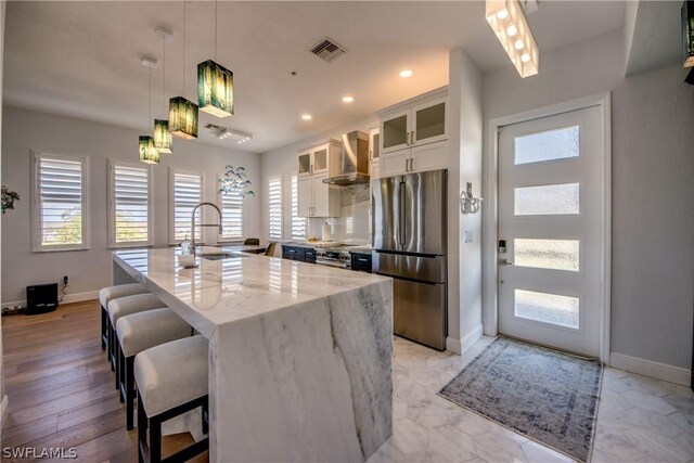 kitchen featuring light wood-type flooring, stainless steel appliances, a kitchen island with sink, wall chimney exhaust hood, and sink