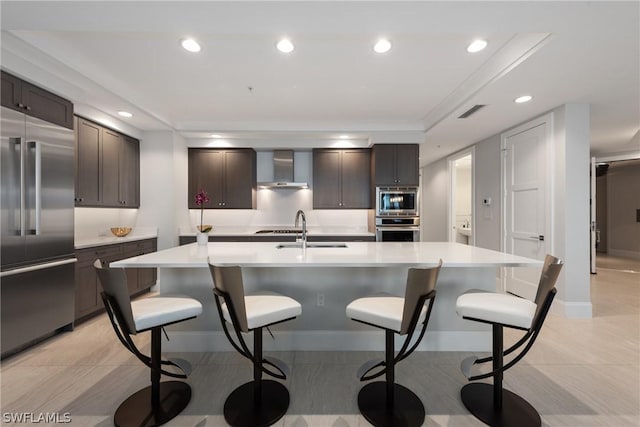 kitchen featuring sink, built in appliances, wall chimney exhaust hood, and an island with sink