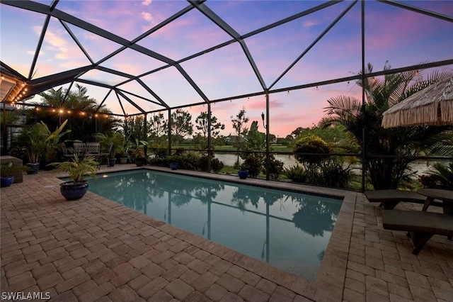 pool at dusk featuring glass enclosure, a water view, and a patio