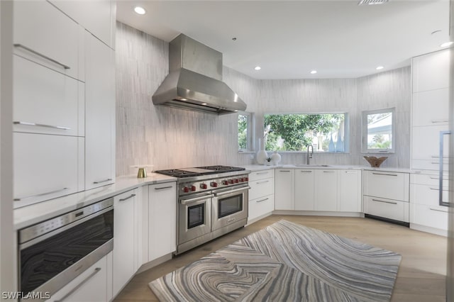 kitchen with wall chimney range hood, oven, white cabinetry, double oven range, and sink