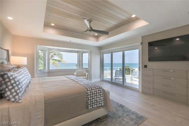 bedroom with ceiling fan, a tray ceiling, a water view, light wood-type flooring, and access to exterior