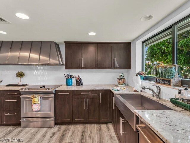 kitchen featuring dark brown cabinets, appliances with stainless steel finishes, light stone counters, light wood-style floors, and wall chimney exhaust hood