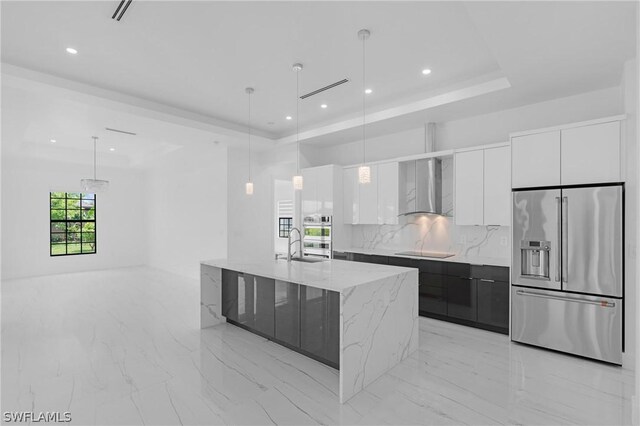 kitchen featuring wall chimney range hood, high end refrigerator, an island with sink, decorative light fixtures, and white cabinets