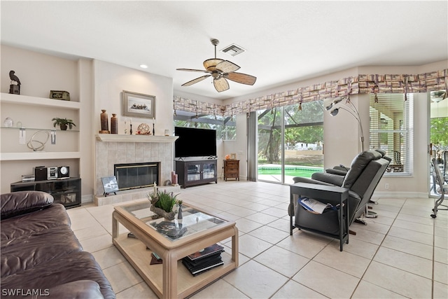 living room featuring built in shelves, a tiled fireplace, ceiling fan, and light tile flooring