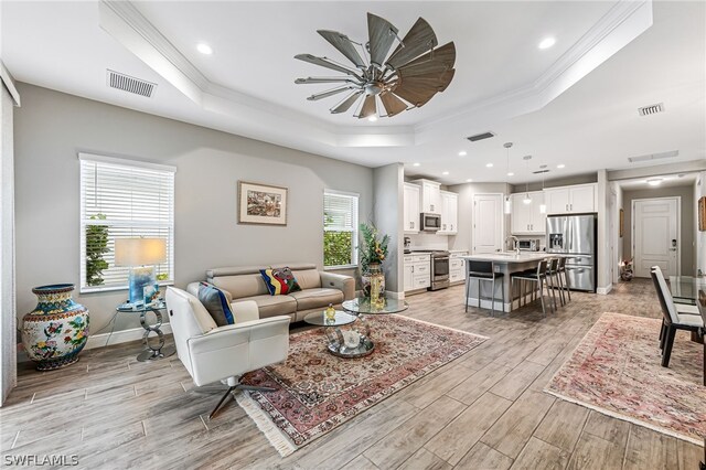 living room featuring a tray ceiling, sink, ornamental molding, and light wood-type flooring