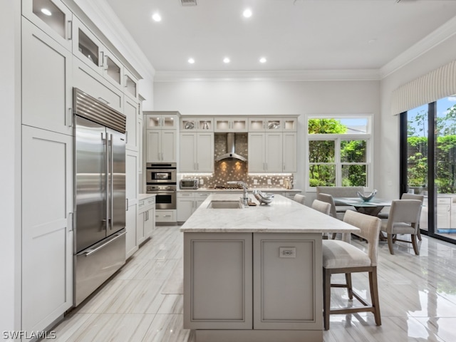 kitchen featuring wall chimney exhaust hood, decorative backsplash, light stone countertops, an island with sink, and appliances with stainless steel finishes