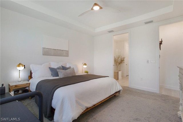 bedroom with visible vents, baseboards, light colored carpet, a tray ceiling, and ensuite bathroom