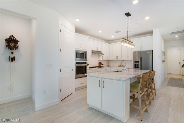 kitchen with sink, appliances with stainless steel finishes, white cabinetry, light stone counters, and a center island with sink