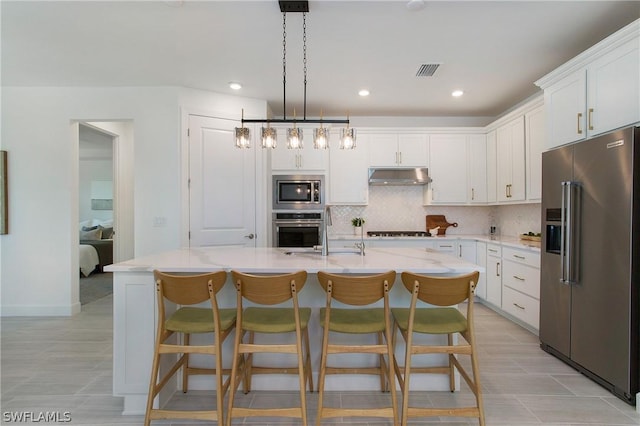 kitchen featuring pendant lighting, appliances with stainless steel finishes, a kitchen island with sink, white cabinetry, and light stone counters